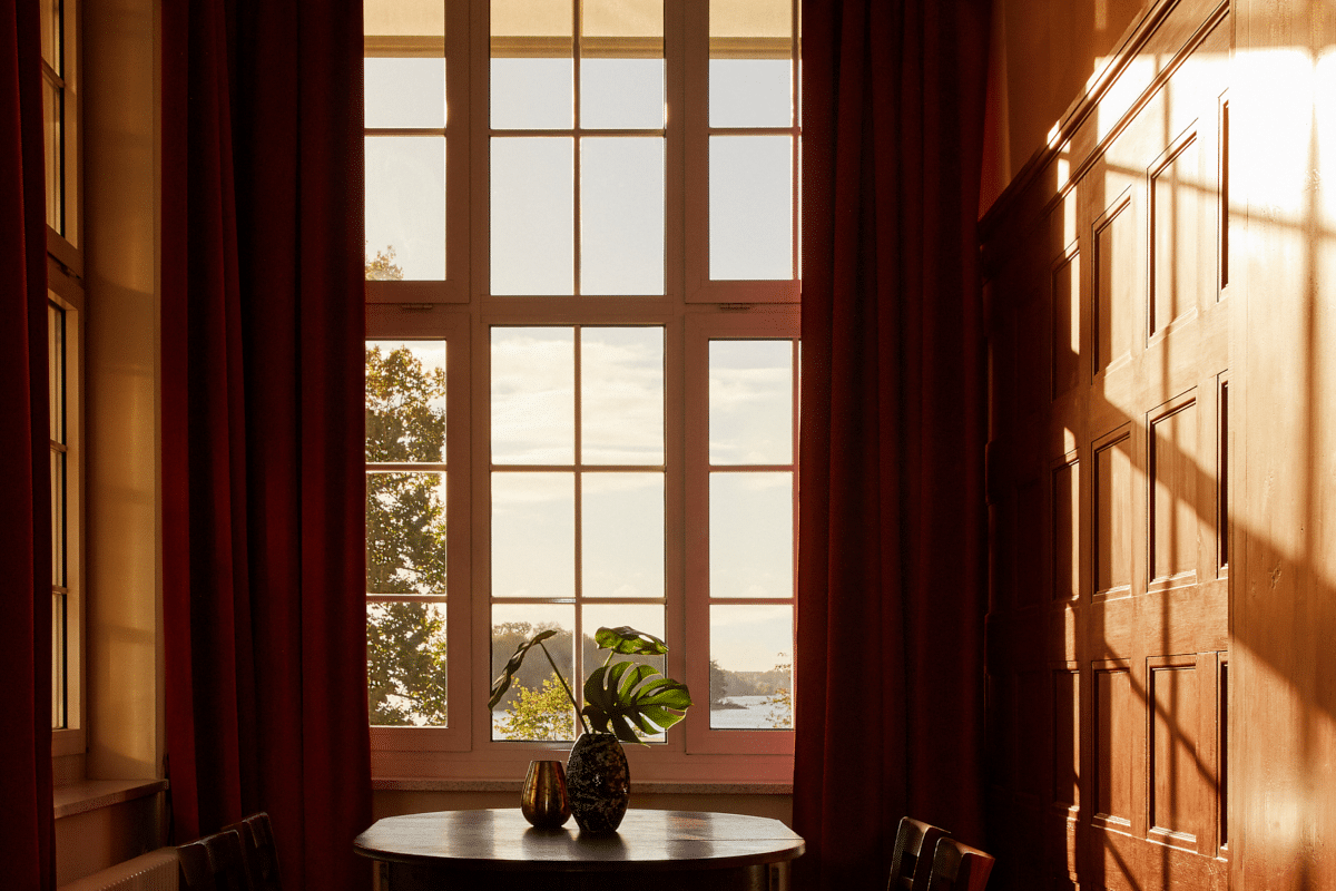 Elegantes Wohnzimmer mit großem Fenster zum See, burgunderroten Vorhängen, Holzpaneelen und rundem Beistelltisch mit Pflanze